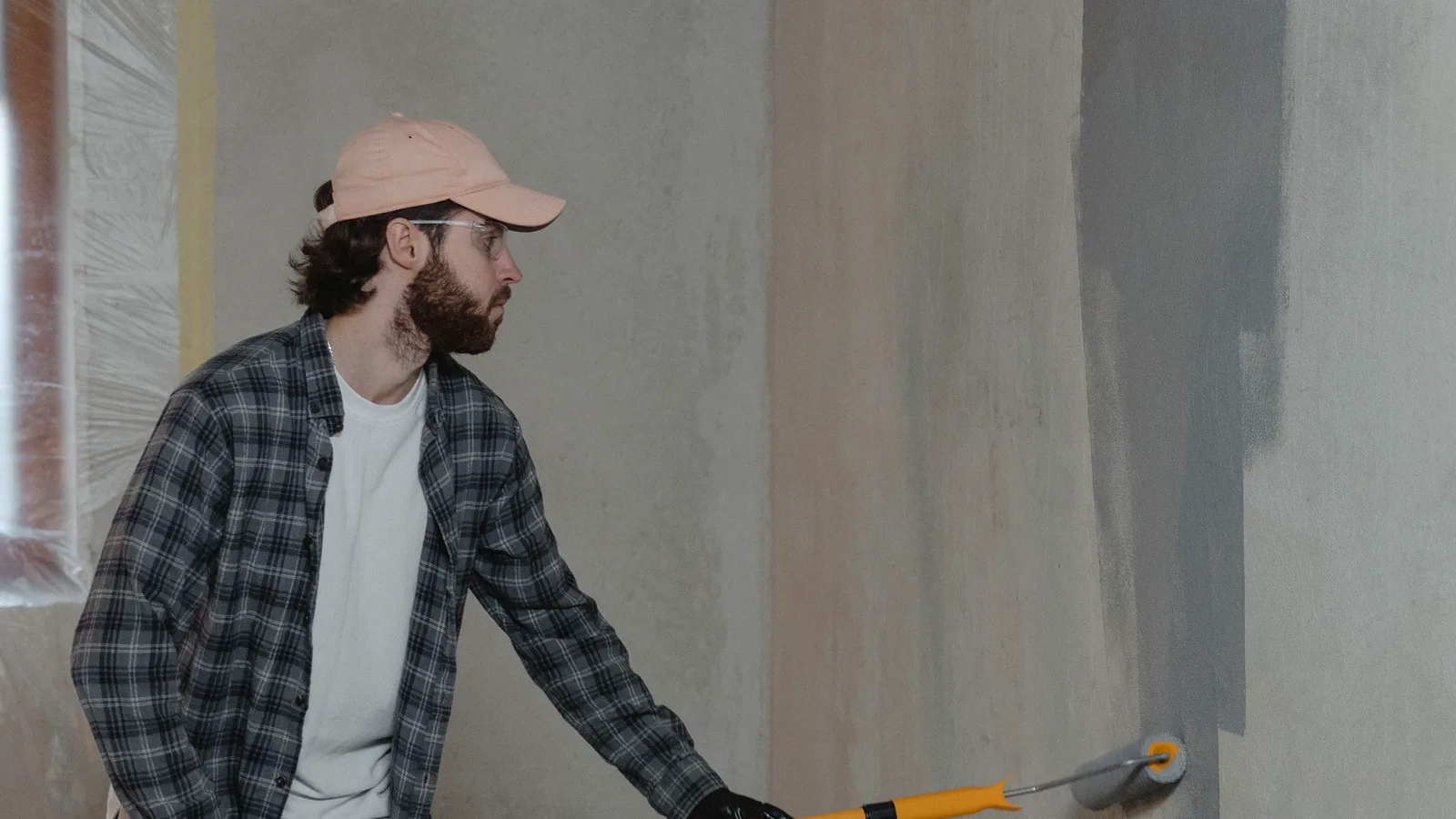 Illustrative image: a painter rolling grey paint onto an interior wall, with the painted and unpainted areas clearly visible side by side.