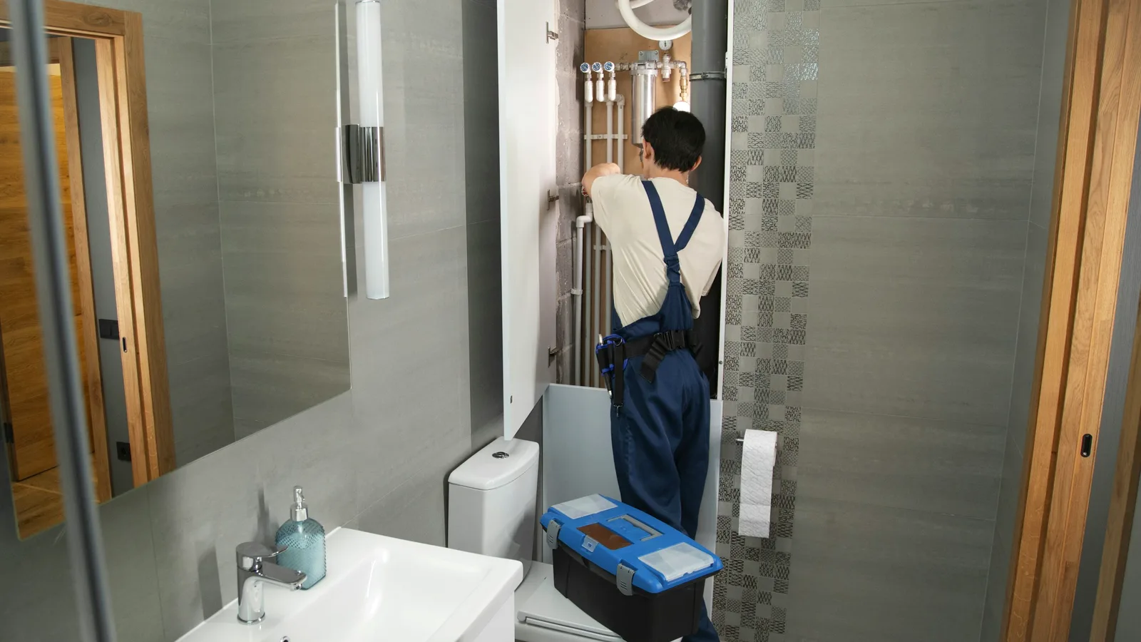 Illustrative image: a plumber working on exposed supply pipes above a bathroom sink and toilet, tool bag in hand.